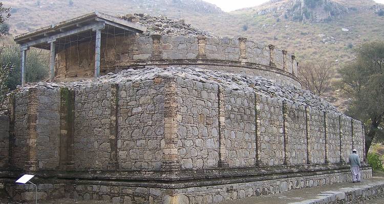 Large circular stone stupa ruin set against a dry hillside; a lone visitor stands at the corner for scale.