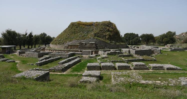 Green meadow surrounds a large grass-covered stupa and stone foundations of the Taxila Buddhist site.