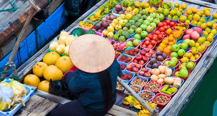 Bovenaanzicht van een bootje op een drijvende markt volgeladen met kleurrijke vruchten terwijl de verkoper in een puntige hoed pagaait.