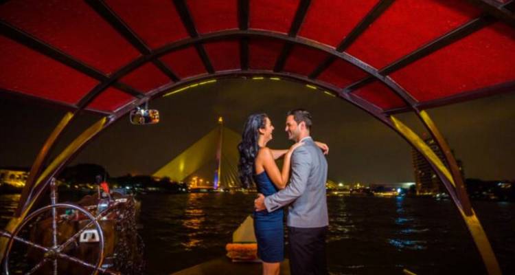 Couple s'embrassant sur un bateau traditionnel la nuit avec les rives illuminées de Bangkok en arrière-plan.