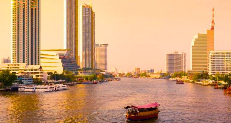 Vue panoramique de la ligne d'horizon des gratte-ciel de Bangkok longeant le fleuve Chao Phraya avec un bateau en bois au premier plan lors d'un coucher de soleil brumeux.