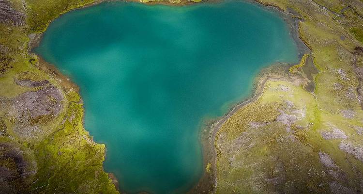 Dronebeeld van een levendige turquoise hartvormige alpinomeer omgeven door mosachtig terrein.