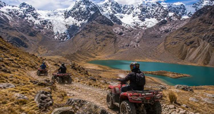 Rijders op rode quads dalen af op een rotsachtig bergpad naar een turquoise alpien meer en besneeuwde toppen.