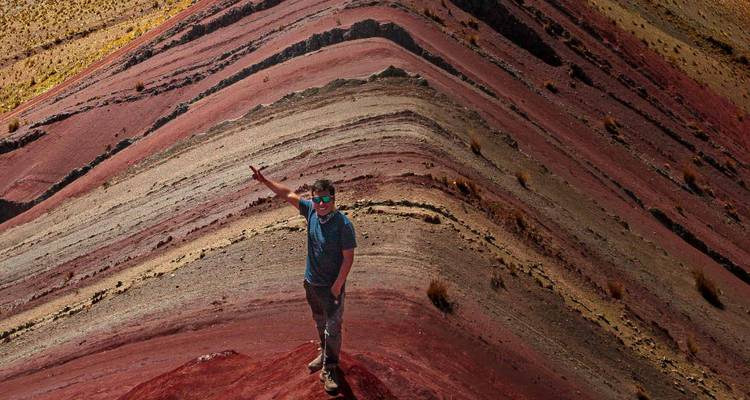 Reiziger poseert bovenop de veelkleurige sedimentlagen van de Rainbow Mountain-bergrug.