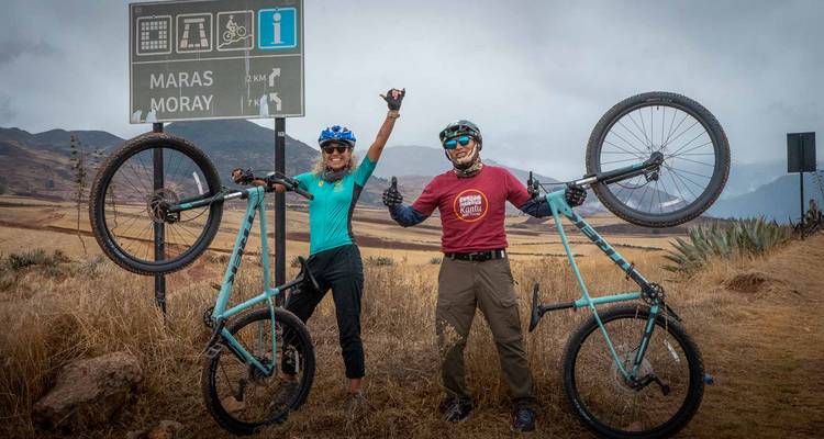 Twee fietsers die triomfantelijk hun mountainbikes rechtop houden naast een wegwijzer die naar Maras en Moray in de hooglanden wijst