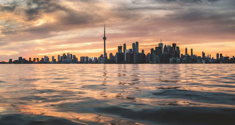 Torontos Skyline im Gegenlicht bei Sonnenuntergang mit farbenfrohen Wolken, die sich im Ontariosee spiegeln.