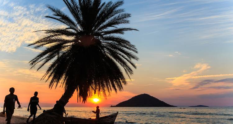 Silhouetten von Menschen und einer geneigten Palme vor einem farbenfrohen Sonnenuntergang über einem ruhigen See und einer Insel