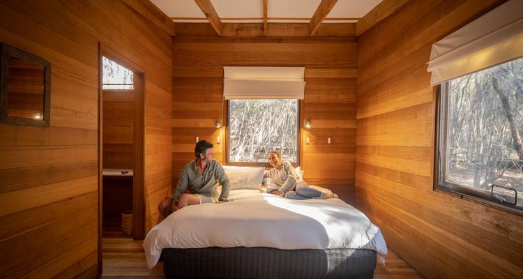 Acogedora habitación de cabaña ecológica con revestimiento de madera, con una cama grande donde una pareja se relaja junto a una ventana con vistas a la luz del bosque.