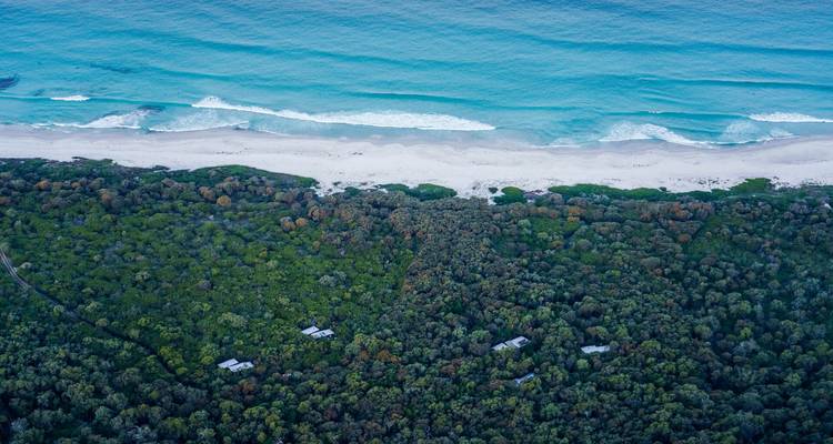 Vista aérea alta de olas de océano turquesa que se encuentran con una playa de arena blanca respaldada por un denso bosque verde con cabañas ecológicas dispersas.