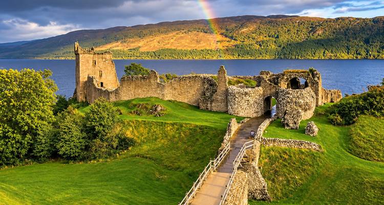 Les ruines du château d'Urquhart sur le Loch Ness illuminées par un arc-en-ciel s'arquant au-dessus de collines dorées à la lumière de fin d'après-midi.