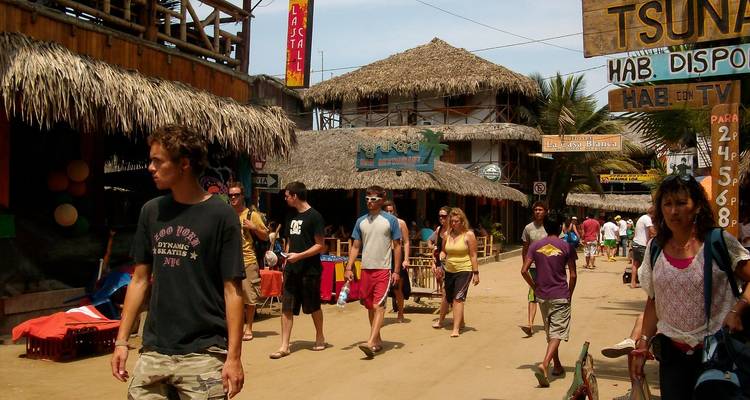 Rue sablonneuse animée d'une petite ville côtière équatorienne réputée pour le surf, bordée de bars en bambou, de commerces et de vacanciers se promenant sur la plage.