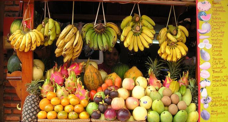 Un étalage de fruits colorés dans la rue, empilé de bananes, de fruits du dragon, d'agrumes et bien plus encore, avec le vendeur souriant partiellement visible dans l'ombre.
