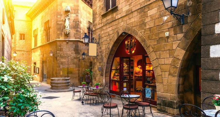 Ruelle européenne pittoresque et étroite avec des bâtiments en pierre, un puits, des tables de café et une porte en arche baignée par la lumière chaude du matin.