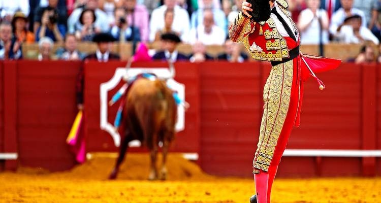 Le matador en costume orné salue la foule dans une arène de corrida aux couleurs vives tandis que le taureau attend en arrière-plan.