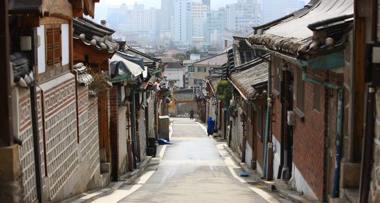 Ruelle étroite bordée de maisons traditionnelles aux toits en tuiles dans un village coréen historique avec les gratte-ciel de la ville moderne en arrière-plan.