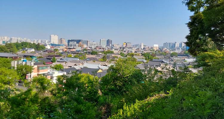 Vue panoramique sur un grand ensemble de toits de hanok coréens traditionnels encadrés par des arbres feuillus, avec un paysage urbain moderne et un ciel bleu clair en arrière-plan.