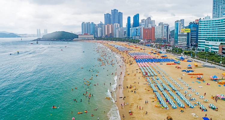 Plage de sable bondée bordée de parasols colorés à côté d'une eau turquoise et en arrière-plan une skyline urbaine moderne avec des immeubles de grande hauteur.