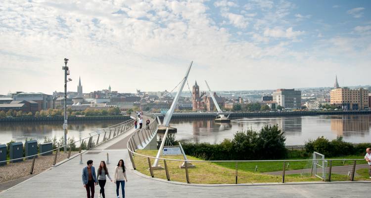 Passerelle moderne en arc au-dessus d'une rivière calme avec la skyline de la ville et des promeneurs profitant d'une matinée ensoleillée.