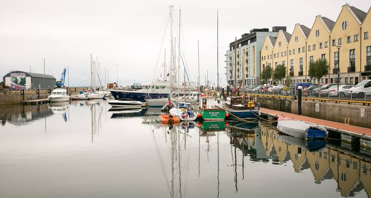 Marina tranquille avec des voiliers et des appartements jaunes en bord de mer reflétés dans les eaux calmes du port.