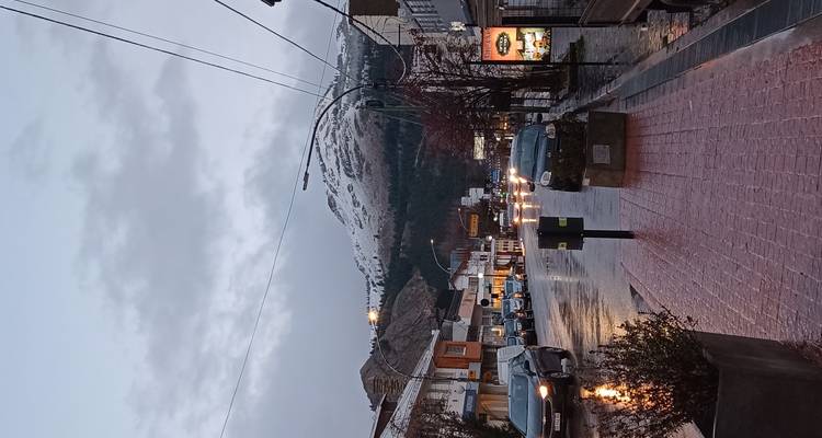 Regenachtige straat van een klein stadje bij schemering met auto's, nat wegdek en sneeuwbergen op de achtergrond.