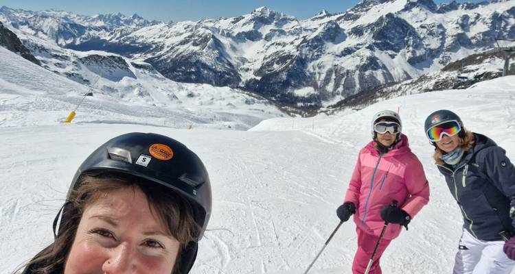 Fröhliche Frauen in Skihelmen machen ein Selfie auf einem verschneiten Abhang mit dramatischen Alpengipfeln im Hintergrund.