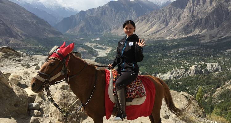 Femme à cheval saluant de la main avec les pics dramatiques enneigés du Karakoram et la vallée verte en contrebas.