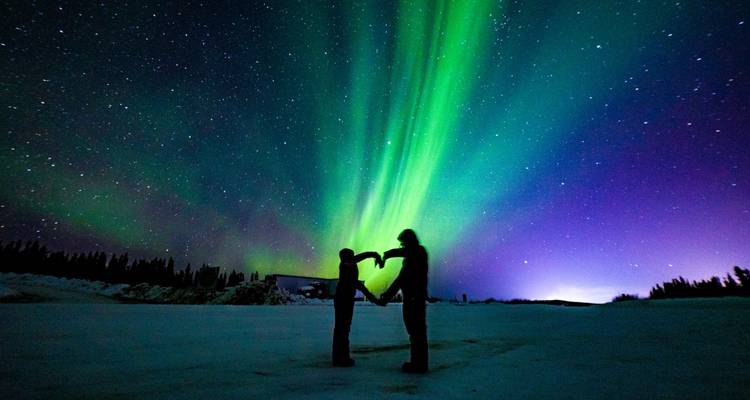 Couple en silhouette formant une forme de coeur sous les aurores boréales d'un vert et d'un violet éclatants au-dessus d'un sol enneigé.