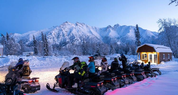 Les motoneigistes font la queue au crépuscule devant de majestueuses montagnes enneigées avec une cabine illuminée à proximité.