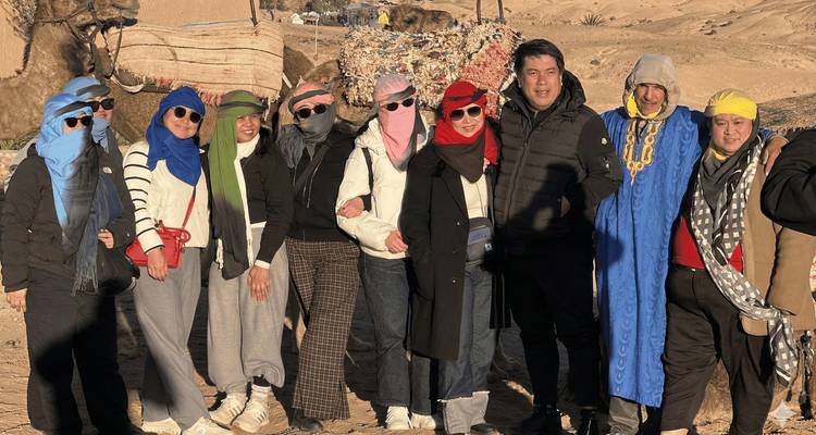 Groupe vêtu de foulards désertiques se tenant près de chameaux et d'une caravane de dunes de sable dans une lumière dorée.