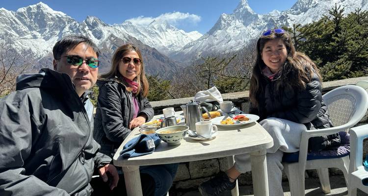 Drie trekkingsliefhebbers genieten van hun ontbijt op een stenen terras met uitzicht op indrukwekkende Himalaya-toppen.