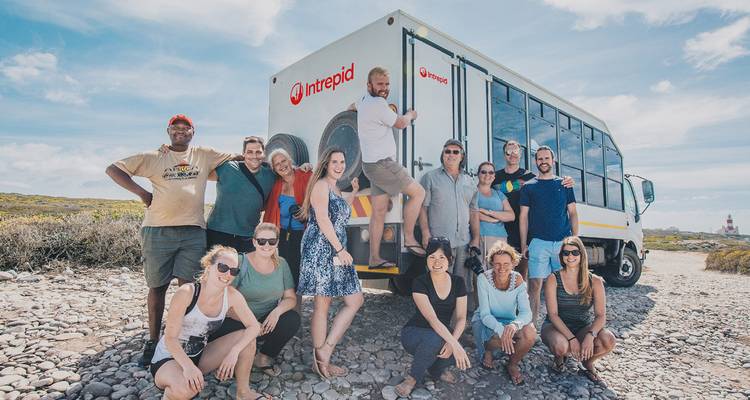 Un grupo de turistas feliz posa junto a un camión Intrepid de viajes por tierra en un camino rocoso junto a la costa.