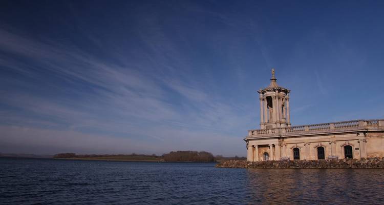 Pabellón de iglesia de piedra histórica se encuentra en la orilla de un enorme lago tranquilo bajo un cielo azul despejado.