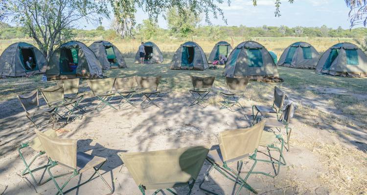 Cirkel van opvouwbare stoelen rond een zandige vuurplaats in een getente bushcamp onder gevlekt schaduw.