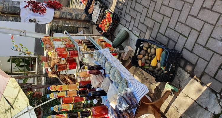 Een marktkraampje in de buitenlucht toont potten met inmaakwerk, flessen, droge waren en verse producten op een kasseienstraat.