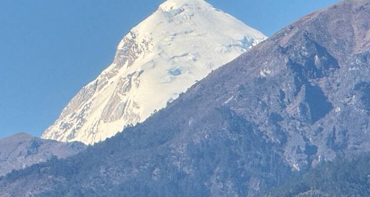 Een sneeuwbedekte Himalayatop steekt scherp af tegen een donkerder beboste bergrug onder heldere hemels.