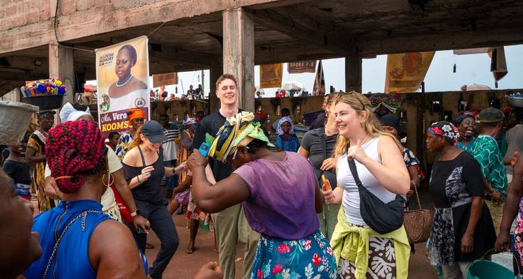 Marché local animé où les voyageurs dansent et interagissent avec les vendeurs sous un toit en béton.