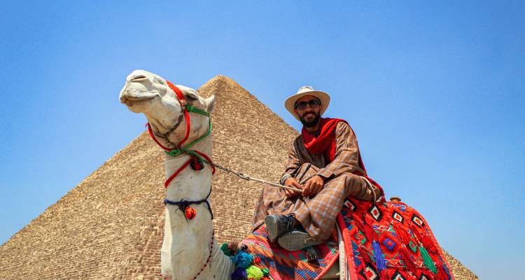 Un homme en tenue traditionnelle est assis sur un chameau décoré devant une pyramide sous un ciel dégagé