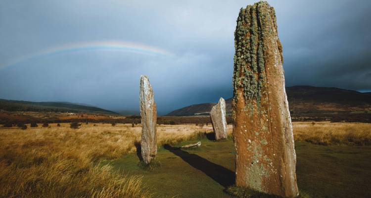 De hautes pierres dressées couvertes de lichen sur une lande ouverte avec un léger arc-en-ciel au-dessus.