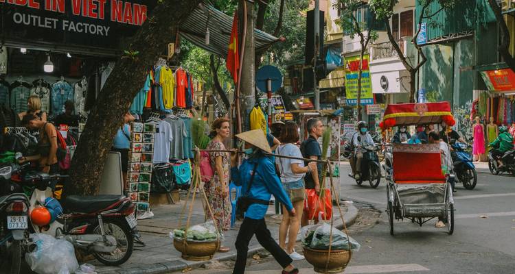 Bustling Hanoi street with vendors, cyclos, and shoppers under colorful shopfronts.