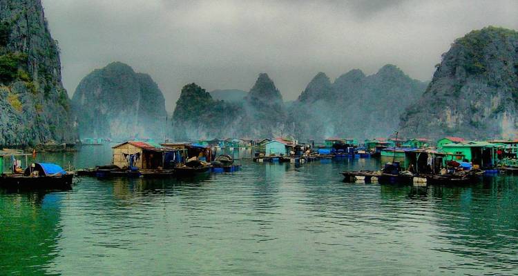 Floating fishing village amid misty limestone peaks of Halong Bay under gray skies.