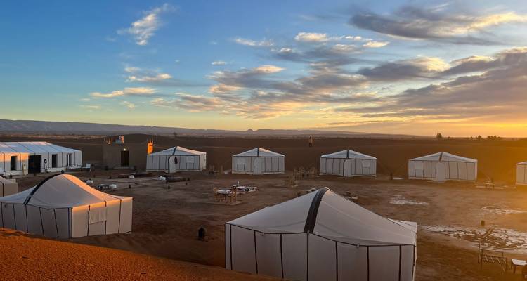 Camp de luxe dans le désert avec des tentes blanches disposées autour d'un foyer ouvert au coucher du soleil.