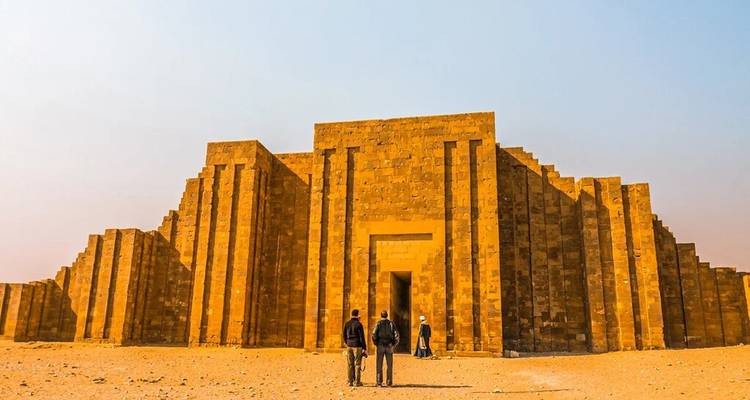 Reizigers naderen de imposante trappentreden van een oud zandstenen complex in de woestijn onder een mistige hemel.