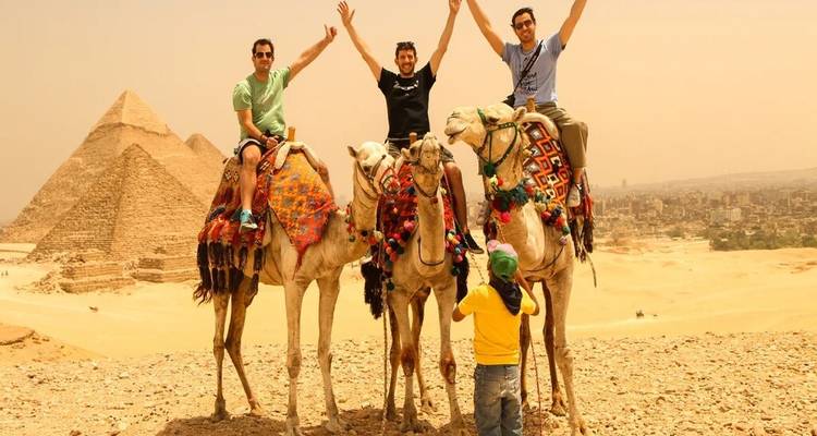 Vier vrienden steken hun armen triomfantelijk in de lucht terwijl ze op kamelen zitten met de piramides van Giza achter hen.