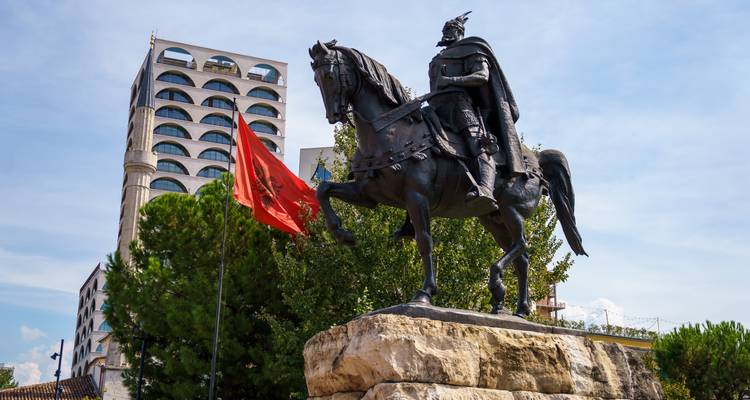 Statue équestre avec le drapeau albanais flottant devant une toile de fond de bâtiments modernes.