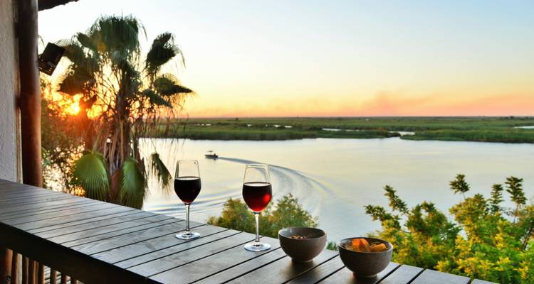 Deux verres de vin rouge sur une terrasse en bois surplombent une rivière au coucher du soleil avec un petit bateau qui laisse un sillage derrière lui.