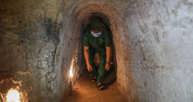 Guide wearing a mask crouches inside a dimly lit narrow tunnel passage.