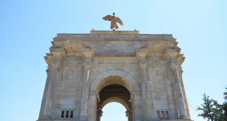 Monument en pierre avec une statue d'ange au sommet d'une arche sur un ciel bleu dégagé.