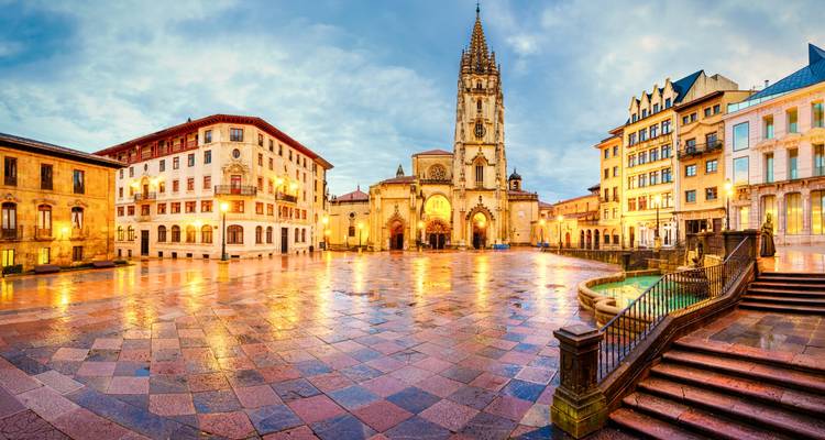 Plaza mojada por la lluvia reflejando luces frente a la Catedral de Oviedo al amanecer.