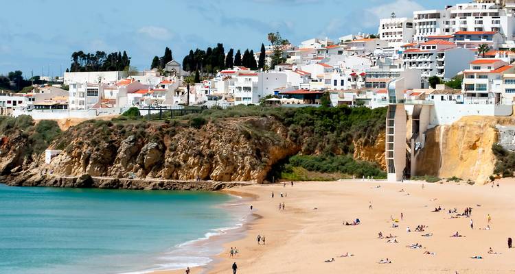 Wide sandy beach below whitewashed hillside houses of Albufeira with sunbathers and turquoise water.