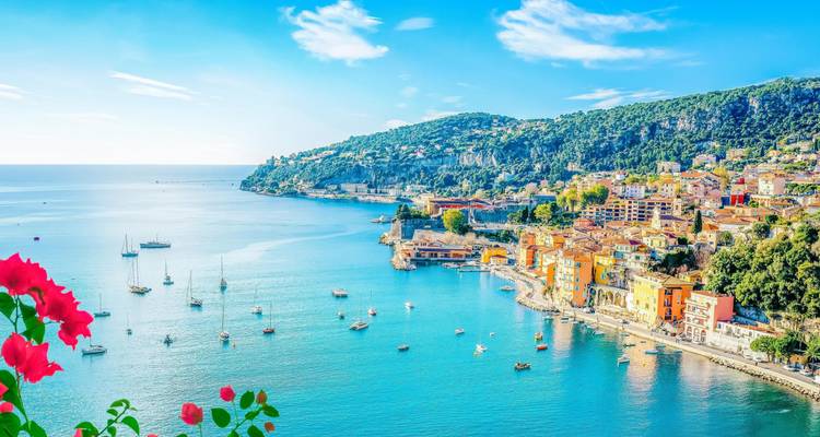 Vue panoramique radieuse sur Villefranche-sur-Mer avec ses maisons aux couleurs pastel, ses yachts et la baie méditerranéenne bleue encadrée de fleurs.